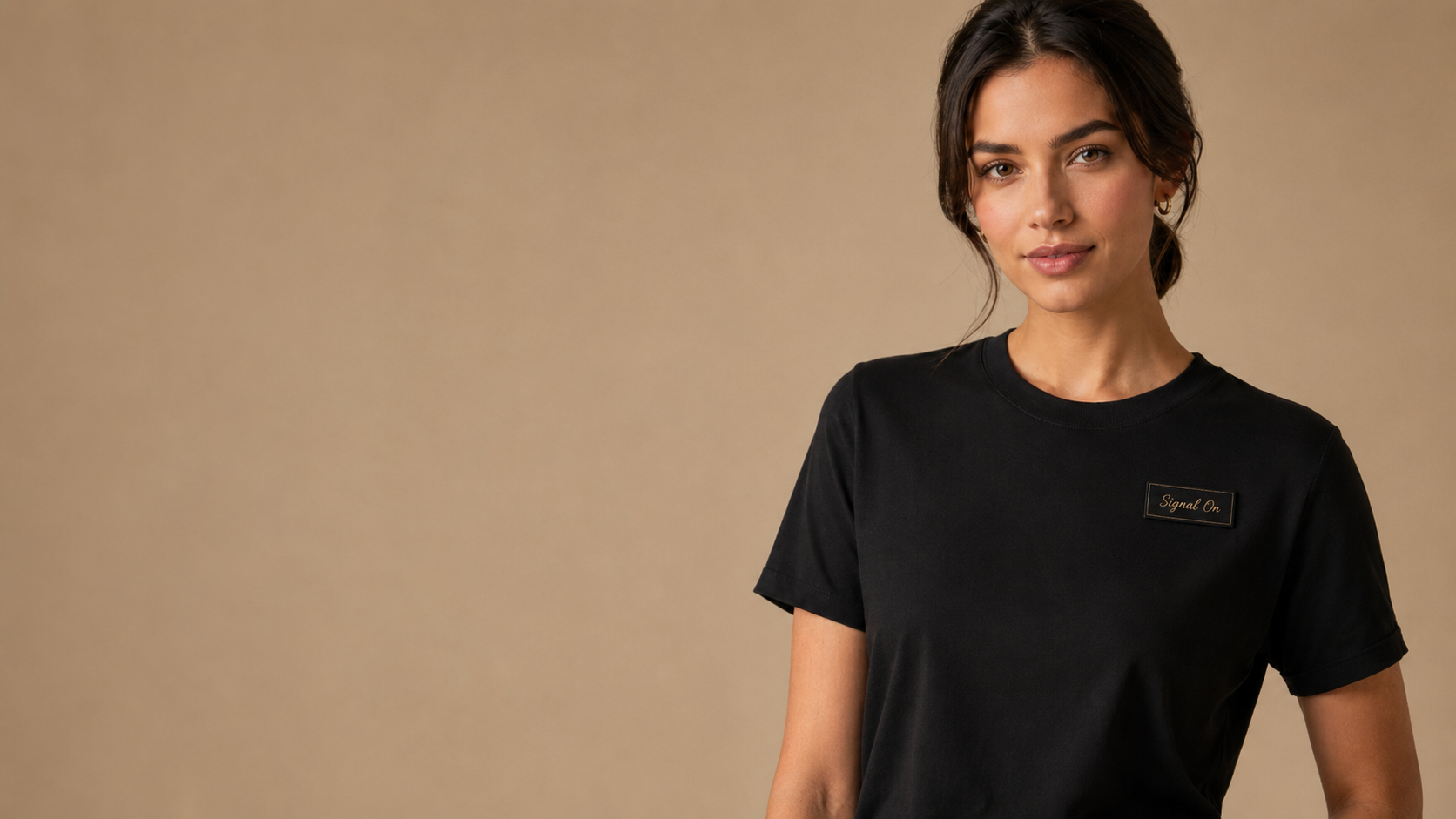 Model wearing a black SendMySignal tee with a small Signal On patch against a warm neutral studio background.